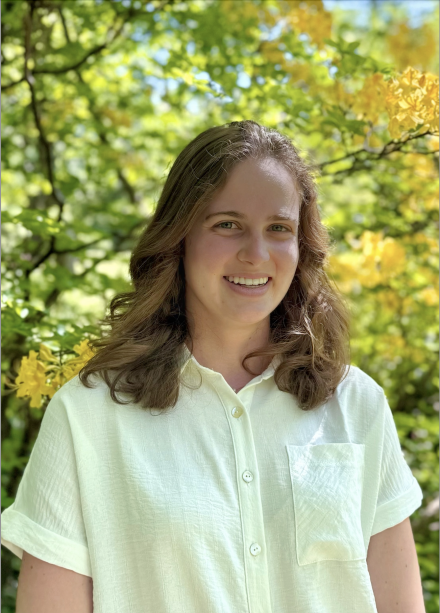 A headshot of Aubrey Spicola '25, wearing a yellow button down shirt in front of a flowering bush.